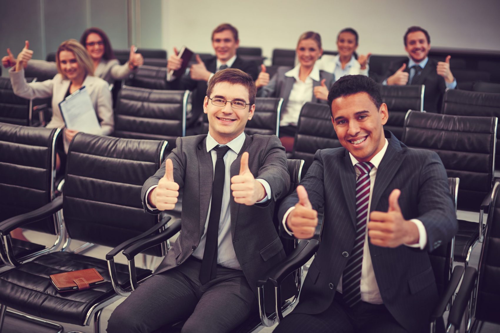 Two happy young businessmen showing thumbs up on background of their colleagues